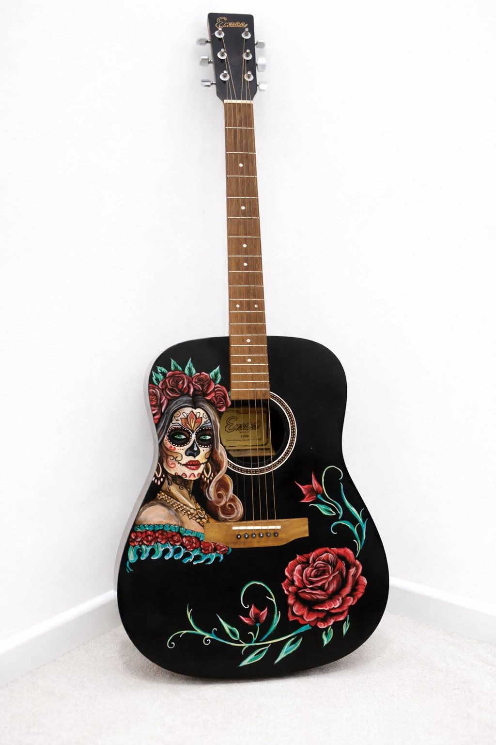 A black acoustic guitar with a Day of the Dead-themed painting, featuring a woman's face and roses, leaning against a whit...